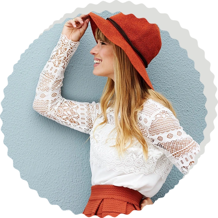 A person wearing a white lace blouse and an orange hat smiles while looking sideways against a light blue background, exuding a sense of zodiac sign fashion.