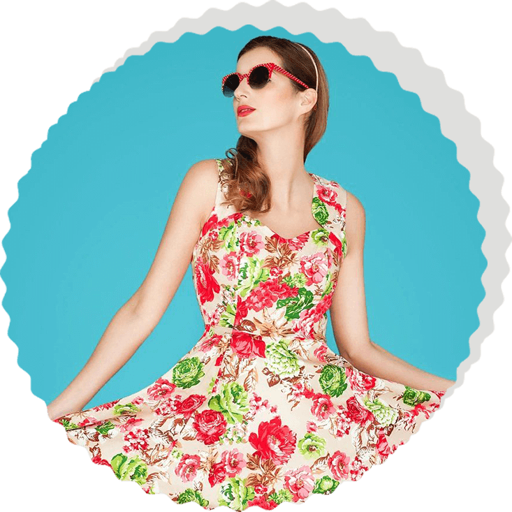 A woman wearing a floral dress and red sunglasses stands against a blue background. She holds out the sides of her dress, showcasing its pattern of red and green flowers that seem as if they were aligned by the stars in an exquisite display of astrology.